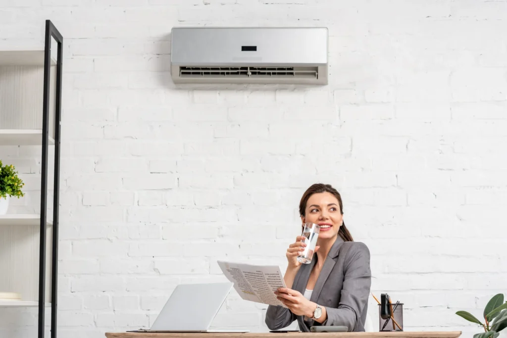 ac companies office worker reviewing a document and drinking from a glass at a desk with a laptop, under a wall-mounted air conditioning unit
