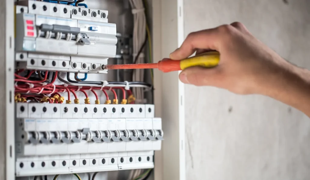a person is using a screwdriver to work on a circuit breaker panel. the panel has numerous white breakers with black switches and red and black wiring.