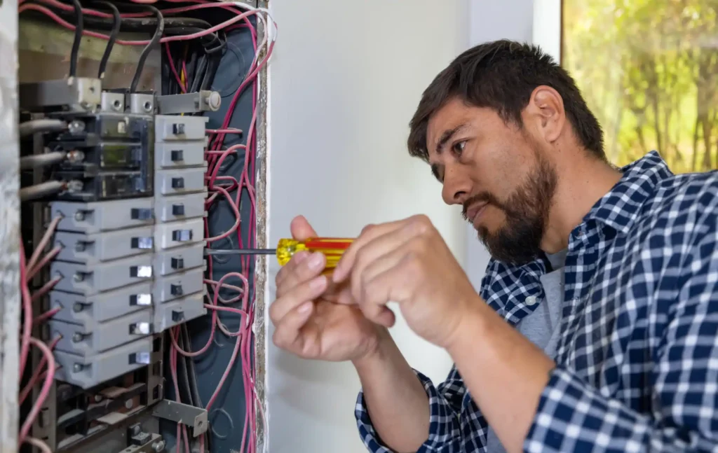 technician in checkered shirt using a screwdriver to service an electrical panel with multiple circuit breakers and wires