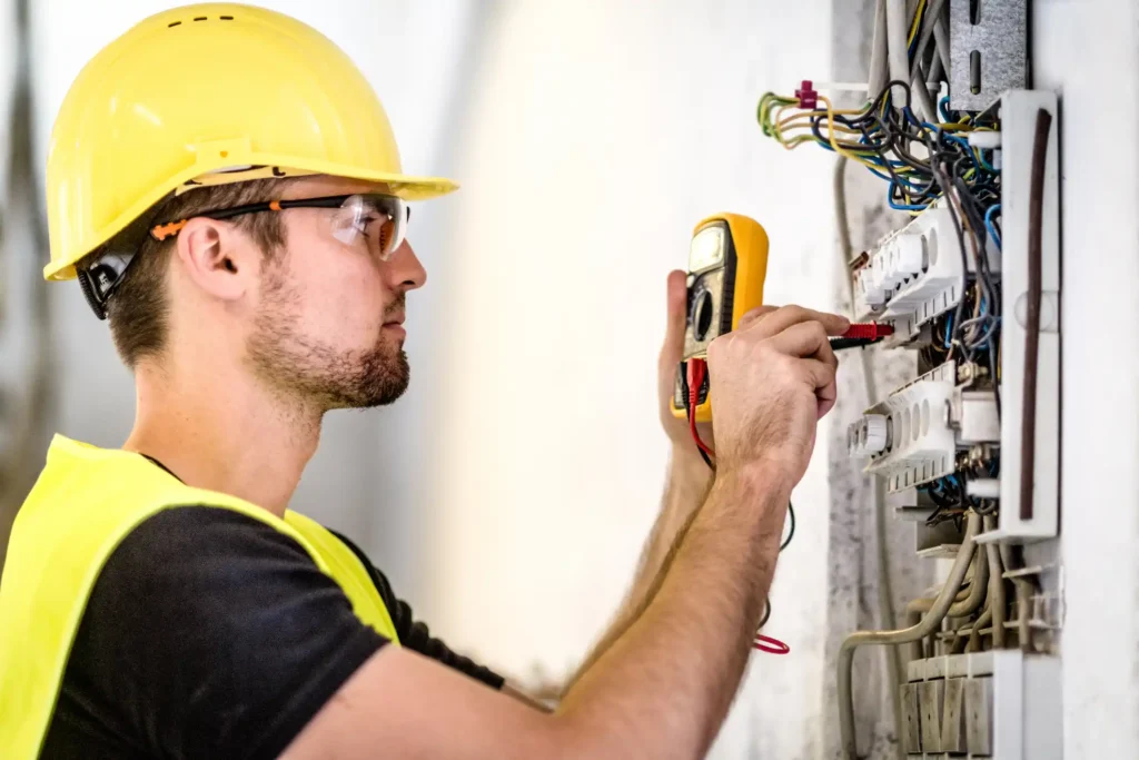 electrical panel installation technician in yellow hard hat and safety vest using an orange multimeter to test electrical connections in a panel with multicolored wires